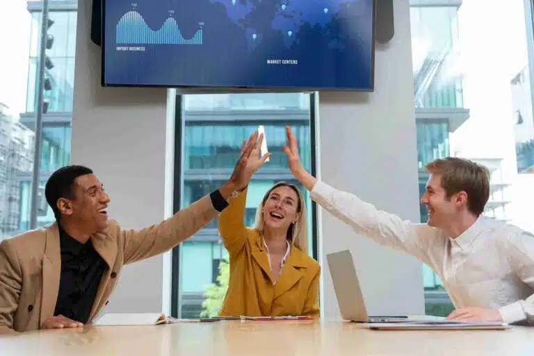 Equipe de trabalho comemorando metas alcançadas em reunião, trocando high five diante de tela com gráficos de desempenho.