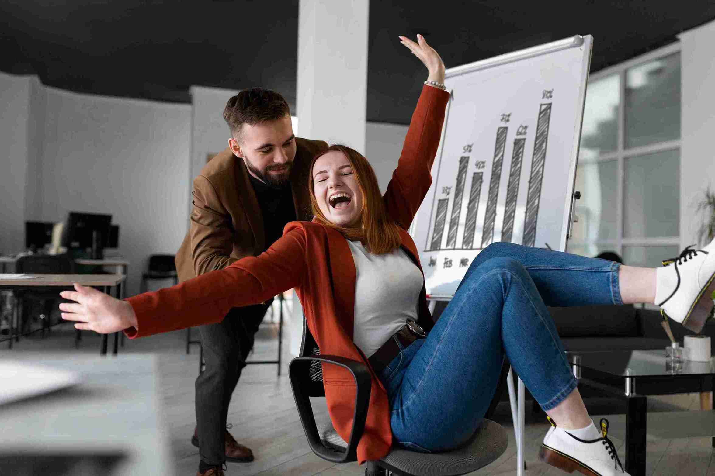 Mulher sorridente sendo empurrada na cadeira de escritório por colega, celebrando bons resultados de trabalho em ambiente corporativo.
